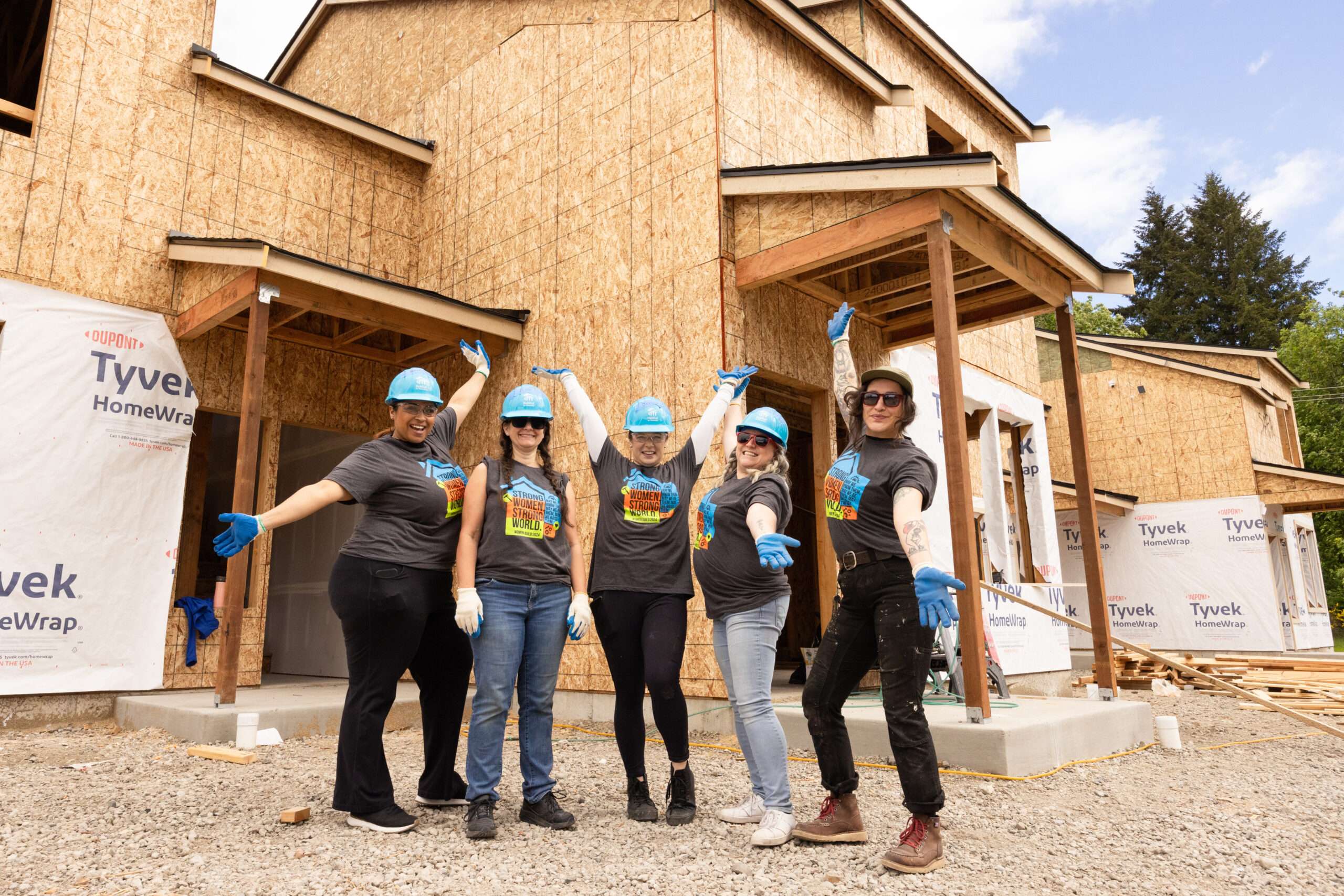 Five women volunteers wearing blue hard hats stand in front of a home under construction.